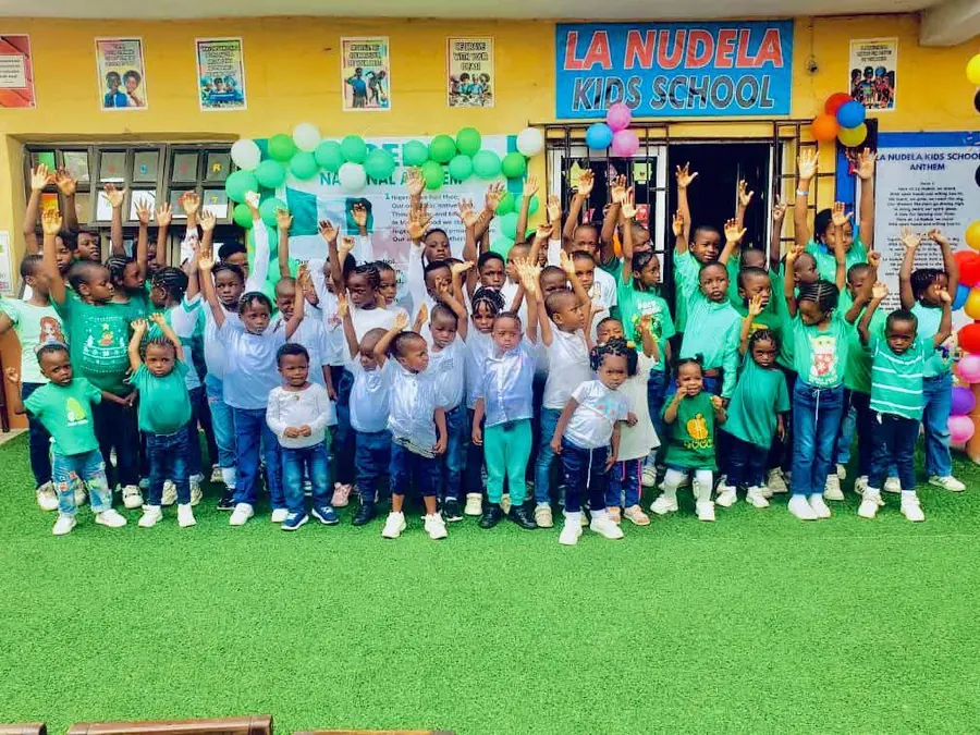 Pupils in green and white raising their hands together outside the school building on National Day